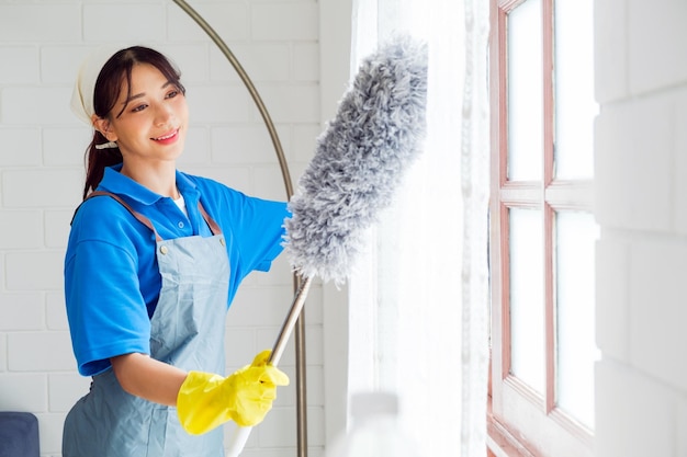 Asian young professional cleaning service woman worker working in the house The girl cleans the curtain and window with a grey feather duster Professional cleaner and chore service concept