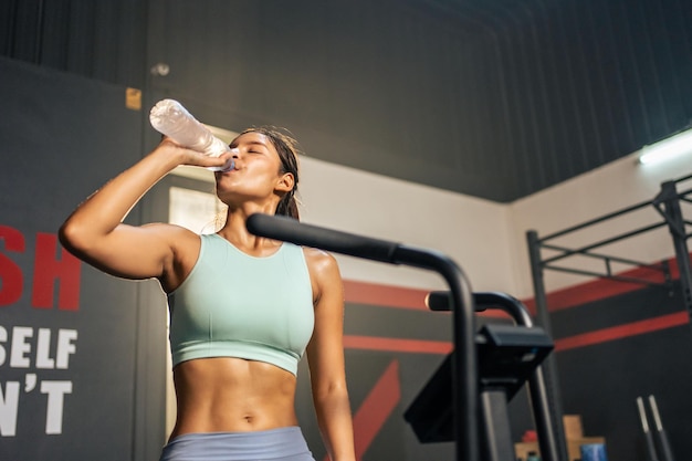 Asian young sportswoman drink a bottle of water after workout in gym Attractive beautiful athlete girl take a break while exercise to maintain muscle for health care and wellbeing at fitness stadium