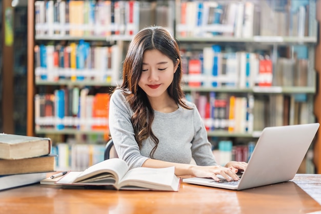 Азиатский молодой студент в повседневном костюме делает домашнее задание и используя технологию ноутбука в библиотеке университета