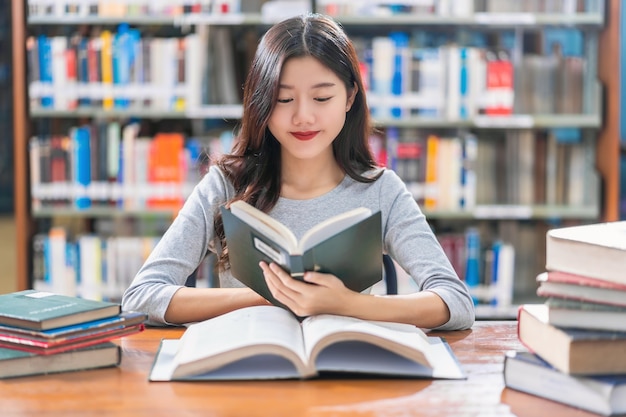 Giovane studente asiatico in abito casual che legge il libro sul tavolo di legno nella biblioteca dell'università