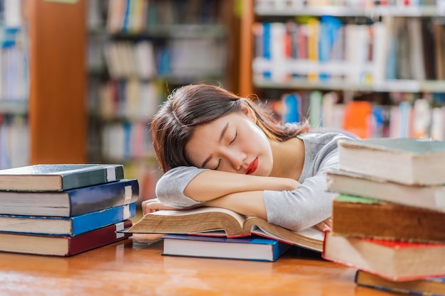 Giovane studente asiatico in abito casual che legge e dorme sul tavolo di legno con vari libri i