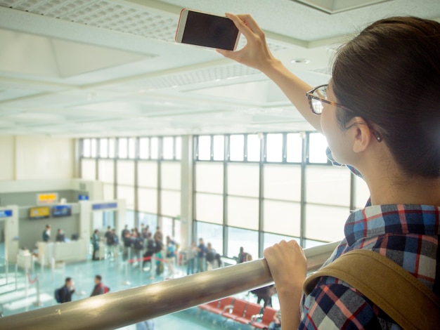 アジアの若い学生が空港に立って、携帯電話を使って自分の写真を撮り、海外での留学旅行の思い出を作ります。