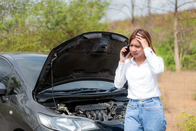 Giovane donna infelice asiatica che parla su un telefono cellulare davanti all'automobile ripartita cappuccio aperto sulla strada campestre che telefona per aiuto.