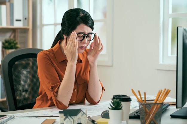アジアの若い女性建築家は、デスクトップコンピュータを使用した後、眼鏡の疲れた目と頭痛を脱いで、現代の職場の机に長時間座っています。眉をひそめている女性デザイナーは目を閉じて疲れ果てた