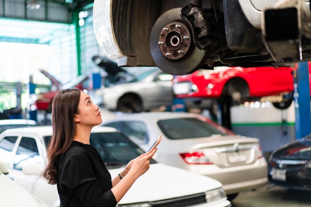 Asian young woman in black shirt examining a vehicle suspension\
and break system in garage close up with copyspace. woman\
inspecting a car on a hydraulic ramp in garage.