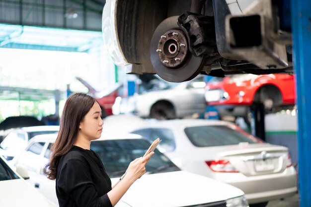 Asian young woman in black shirt examining a vehicle suspension\
and break system in garage close up with copyspace. woman\
inspecting a car on a hydraulic ramp in garage.