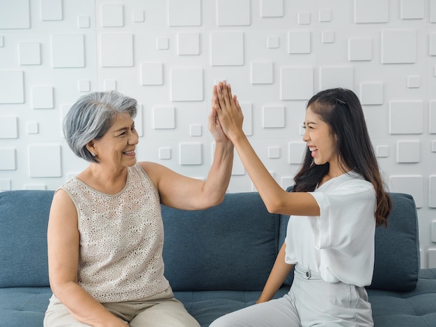 Asian young woman cheerful daughter giving high five to her mom old senior female sitting on grey sofa seat in white room Mother and daughter celebrating victory win together clapping hands