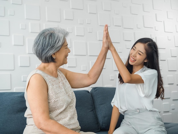 写真 白い部屋で灰色のソファの座席に座っている彼女のお母さん古い年配の女性にハイタッチを与えるアジアの若い女性の陽気な娘母と娘が勝利を祝って一緒に手をたたく