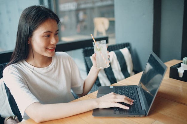 La giovane donna asiatica beve la soda della limonata e usa il computer portatile al caffè all'aperto