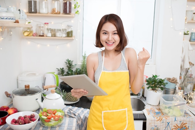 Азиатская молодая женщина ест здоровую пищу и готовит на кухне с помощью планшетного компьютера