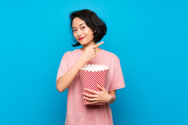 Asian young woman eating popcorns pointing to the side to present a product