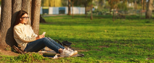 Asian young woman enjoys a peaceful afternoon in the park sitting against a tree and checking her