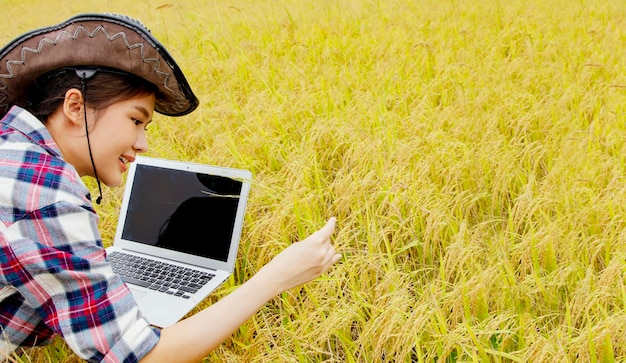Фото Азиатская молодая женщина-фермер в ковбойской шляпе проверяет зерно на зрелом поле с помощью современного ноутбука.