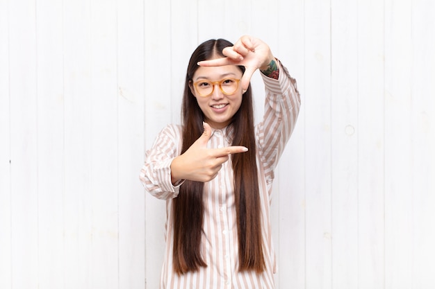 Giovane donna asiatica che si sente felice, amichevole e positiva, sorridente e che fa un ritratto o una cornice con le mani