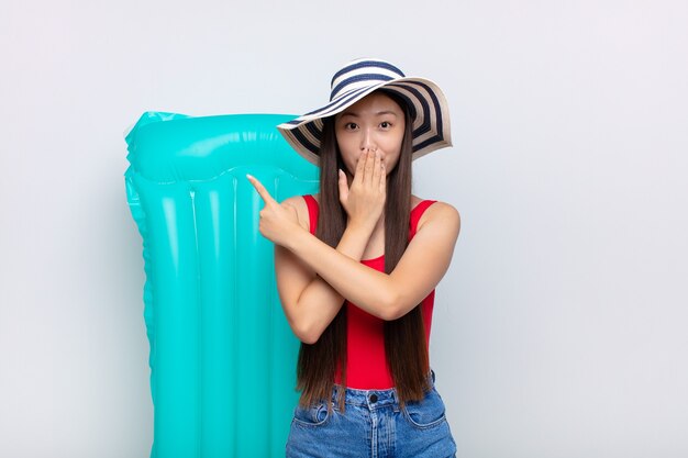 Asian young woman feeling happy, shocked and surprised, covering mouth with hand and pointing to lateral copy space. summer concept