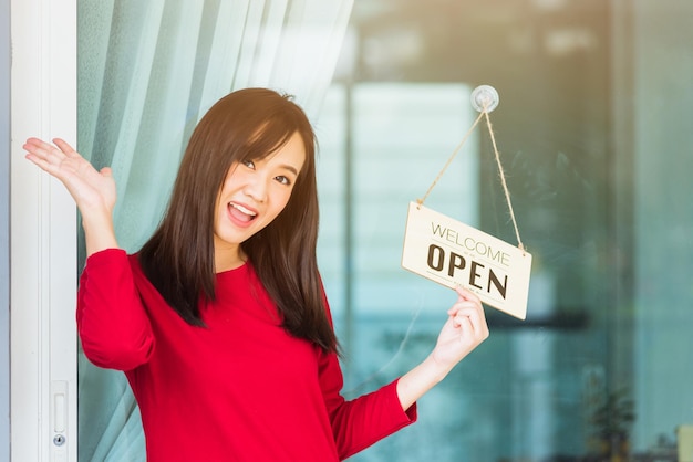 아시아 젊은 여성은 유리문 앞 상점에 걸려 있는 목재 보드 라벨 "WELCOME OPEN"을 보고 기뻐하고 웃고 있으며, 코로나바이러스 대유행 질병 이후 사업이 재개되어 새로운 정상으로 돌아왔습니다.