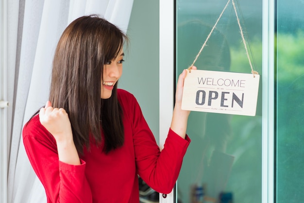 Asian young woman glad and smiling she notice sign wood board label "WELCOME OPEN" hanging through glass door front shop, Business turning open after coronavirus pandemic disease, back to new normal