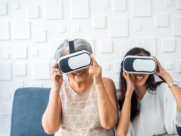 Asian young woman happy daughter and her mother old senior
female wearing vr glasses enjoy 3d game online together in white
room mom and daughter with reality virtual technology lifestyle
concept