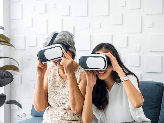 Asian young woman happy daughter and her mother old senior
female wearing vr glasses enjoy 3d game online together in white
room mom and daughter with reality virtual technology lifestyle
concept