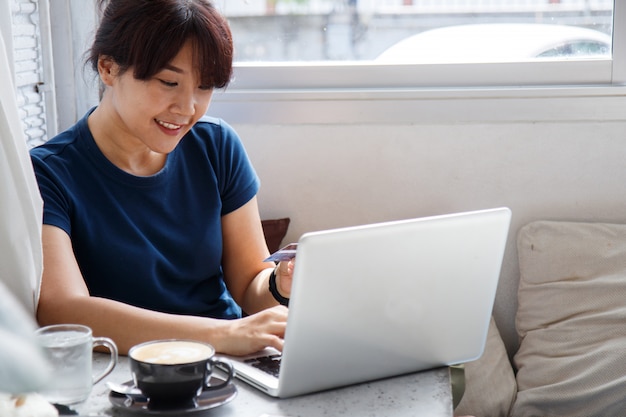 Photo asian young woman holding credit card mockup and using laptop computer while sitting in cafe.