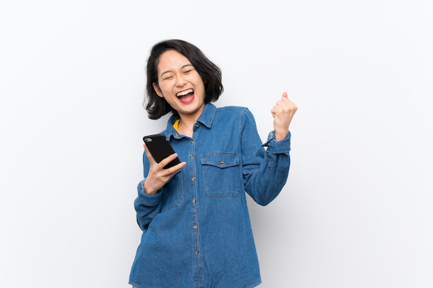 Asian young woman over isolated white wall with phone in victory position