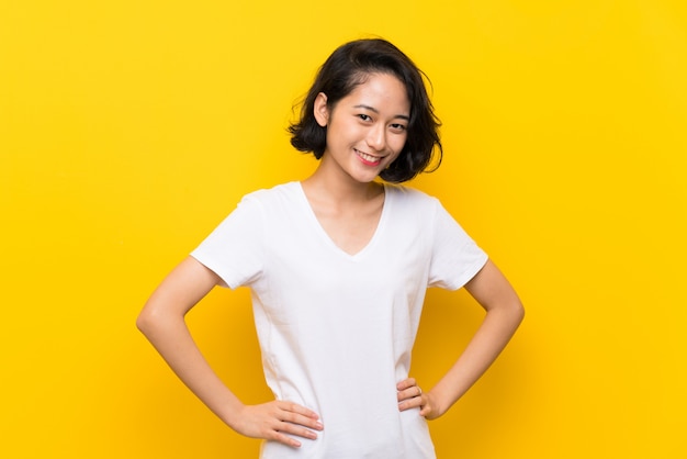 Asian young woman over isolated yellow wall posing with arms at hip and smiling