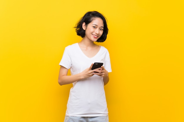 Азиатская молодая женщина над изолированной желтой стеной, отправив сообщение с мобильного телефона