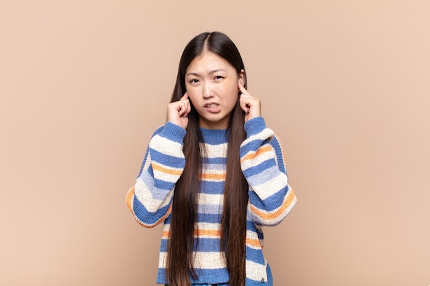 Asian young woman looking angry, stressed and annoyed, covering both ears to a deafening noise, sound or loud music