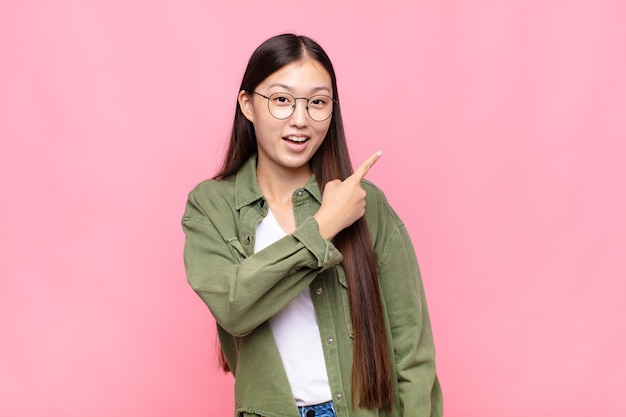 Asian young woman looking excited and surprised pointing to the side and upwards to copy space