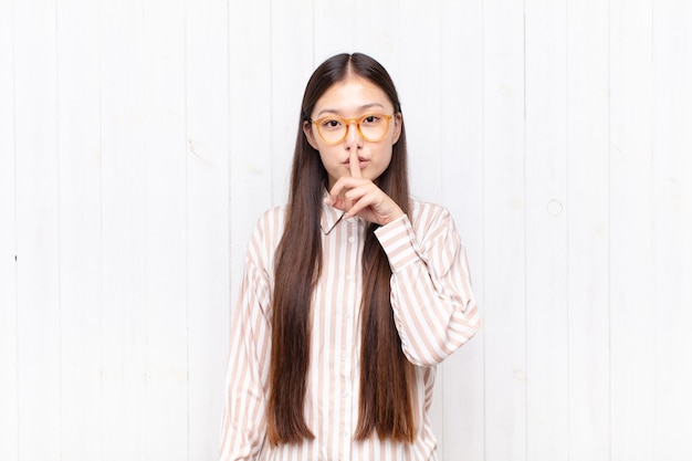 Asian young woman looking serious and cross with finger pressed to lips demanding silence or quiet, keeping a secret