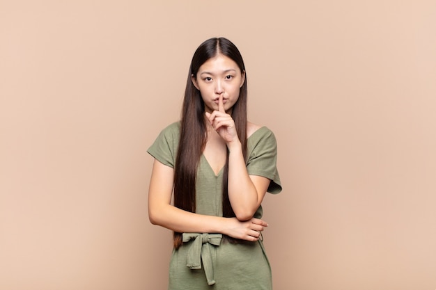 Asian young woman looking serious and cross with finger pressed to lips demanding silence or quiet, keeping a secret