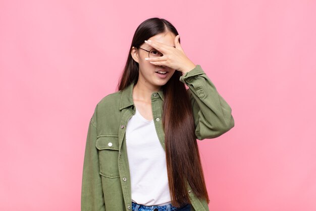 Asian young woman looking shocked, scared or terrified, covering face with hand and peeking between fingers