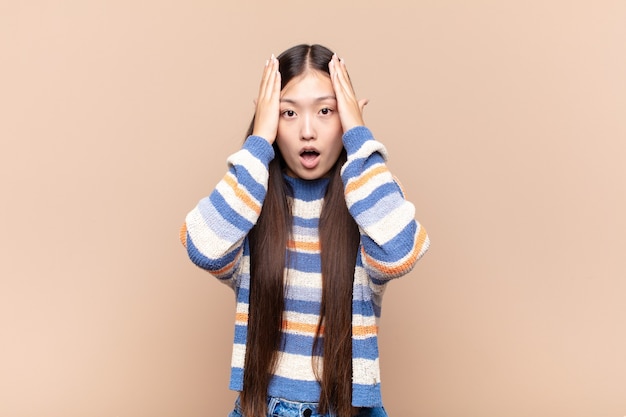 Giovane donna asiatica che sembra spiacevolmente scioccata, spaventata o preoccupata, con la bocca spalancata e coprendo entrambe le orecchie con le mani