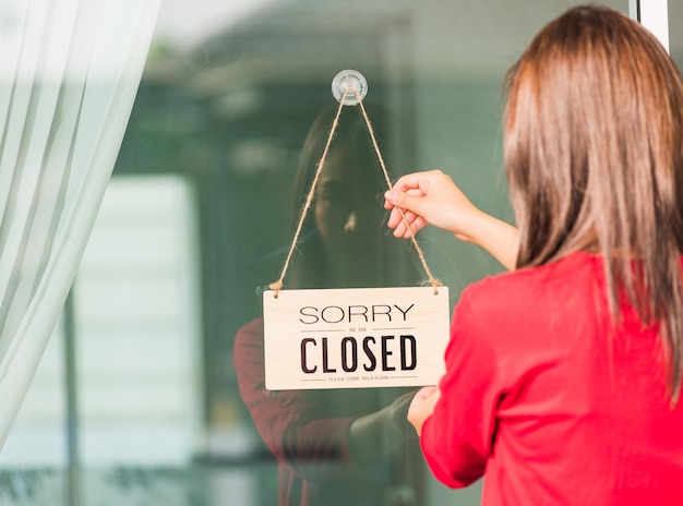 アジアの若い女性の看板木板ラベル「SORRY WE ARE CLOSED PLEASE COME BACK AGAIN」がガラスのドアの前の店にぶら下がり、コロナウイルスのパンデミック病のコンセプトで営業が終了