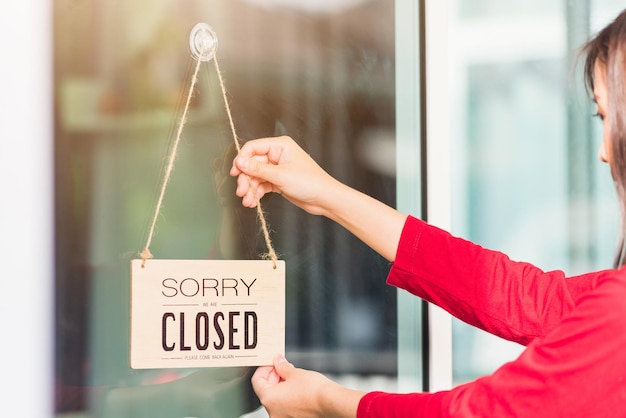 아시아 젊은 여성 공지 표지판 "SORRY WE ARE CLOSED PLEASE COME BACK AGAIN" 유리문 앞 가게에 걸려 있는 나무 판자 라벨