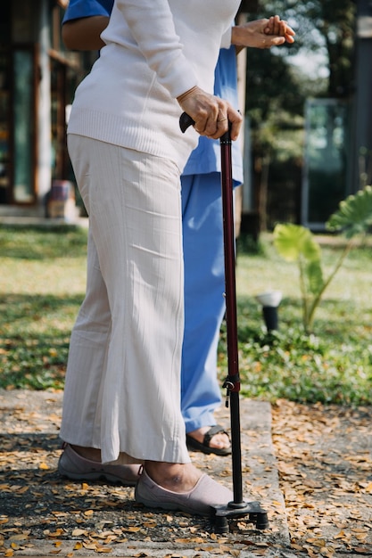 Asian young woman nurse at nursing home take care disabled senior man Caregiver doctor serve physical therapy for older elderly patient to exercise and practice walking on walker or cane at backyard