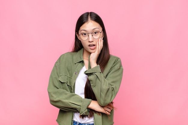 Asian young woman open-mouthed in shock and disbelief, with hand on cheek and arm crossed, feeling stupefied and amazed