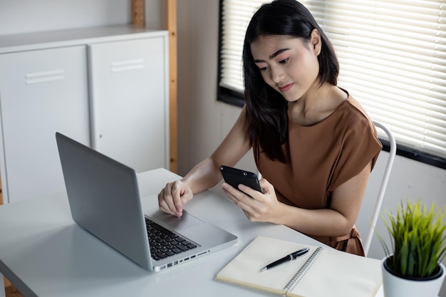 Asian young woman playing social online on mobile with laptop is on table by happy smile in work room at home. Happy woman playing online internet on smartphone.