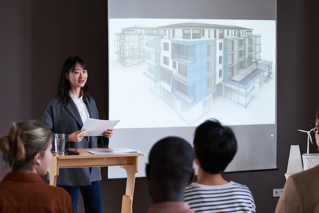 Asian young woman presenting report while standing near the big monitor with blueprint of building and people listening to her