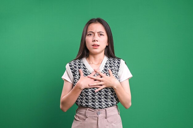 Asian young woman shocked and surprised while holding chest with hands over green background