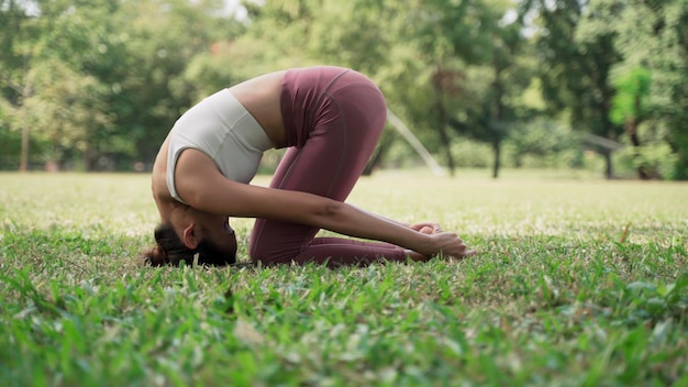 Asian young woman sitting on the grass practicing yoga in the\
cow pose position in city park with the big trees background\
rearview of female practicing yoga outdoors on a sunny day