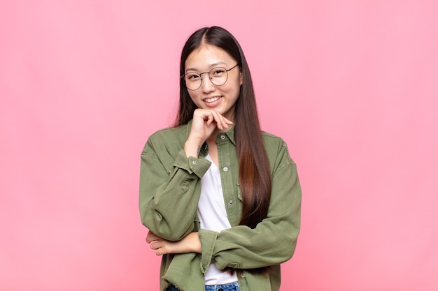 Asian young woman smiling, enjoying life, feeling happy, friendly, satisfied and carefree with hand on chin