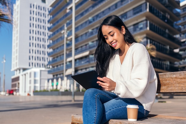 コーヒーの横に彼女のタブレットを使用してベンチに座って笑っているアジアの若い女性