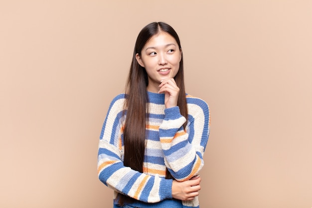 Asian young woman smiling with a happy, confident expression with hand on chin, wondering and looking to the side