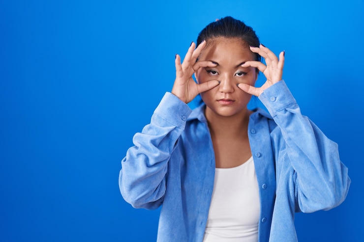 Photo asian young woman standing over blue background trying to open eyes with fingers sleepy and tired for morning fatigue