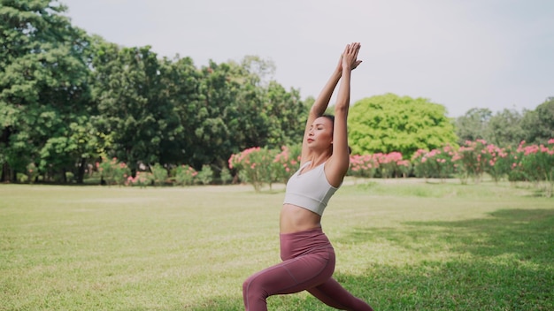 Asian young woman standing on the grass practicing yoga in the Warrior Pose position in city park with the big trees background Rearview of female practicing yoga outdoors on a sunny day