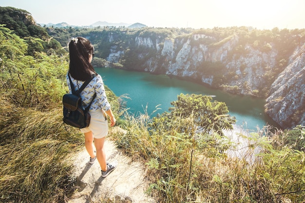 孤独に立って、湖と木と岩山で何かを探しているアジアの若い女性。