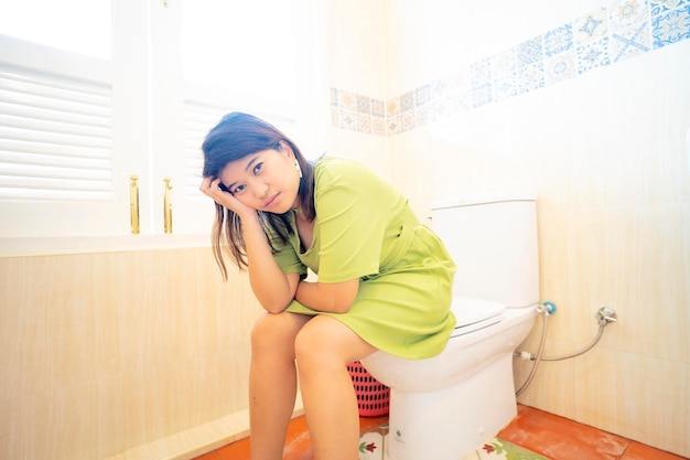 Asian young woman suffering from constipation on toilet bowl at home sitting on toilet defecating