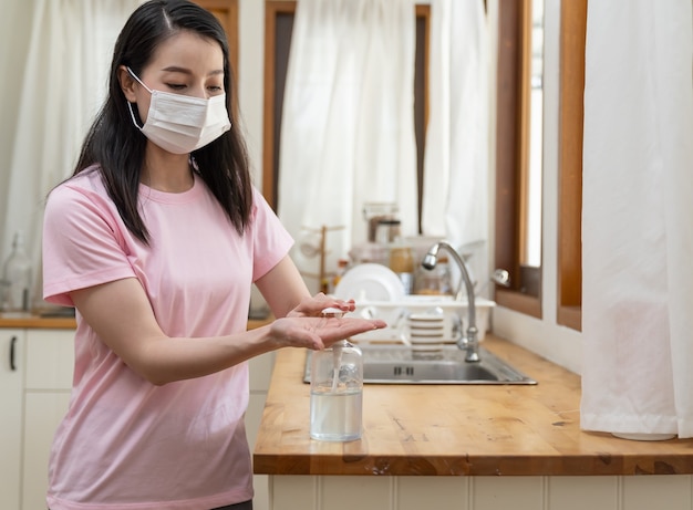 얼굴 마스크 또는 집에서 일하는 보호 마스크를 착용하고 코로나 바이러스 또는 Covid 19 발병 동안 소독제 젤로 손을 청소하는 아시아 젊은 여성.