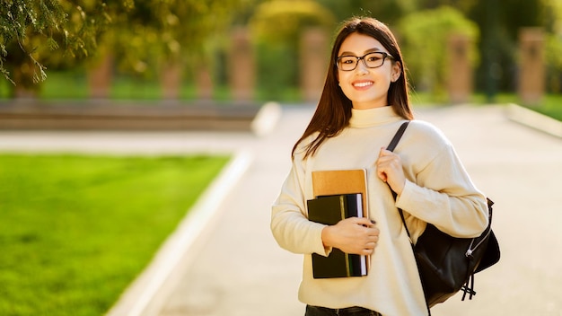 Asian young woman with glasses is walking on a college campus during the morning holding books and a